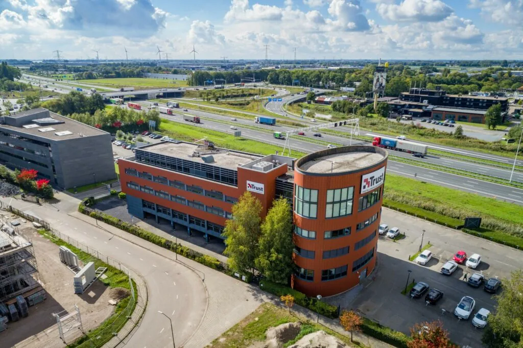 Luchtfoto van het kantoorgebouw van Trust aan de Laan van Barcelona, gelegen naast een snelweg met verkeer en omringd door zakelijke panden en landschap.